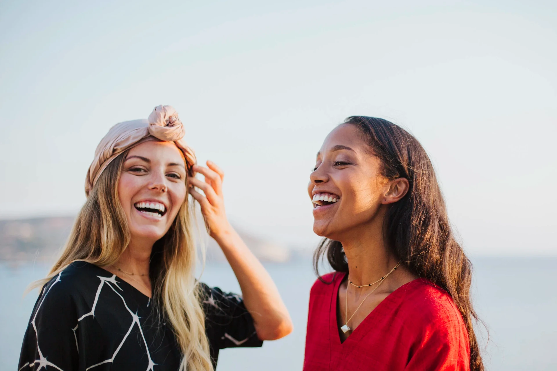 Two models laughing by the sea in a black print kaftan and red linen dress