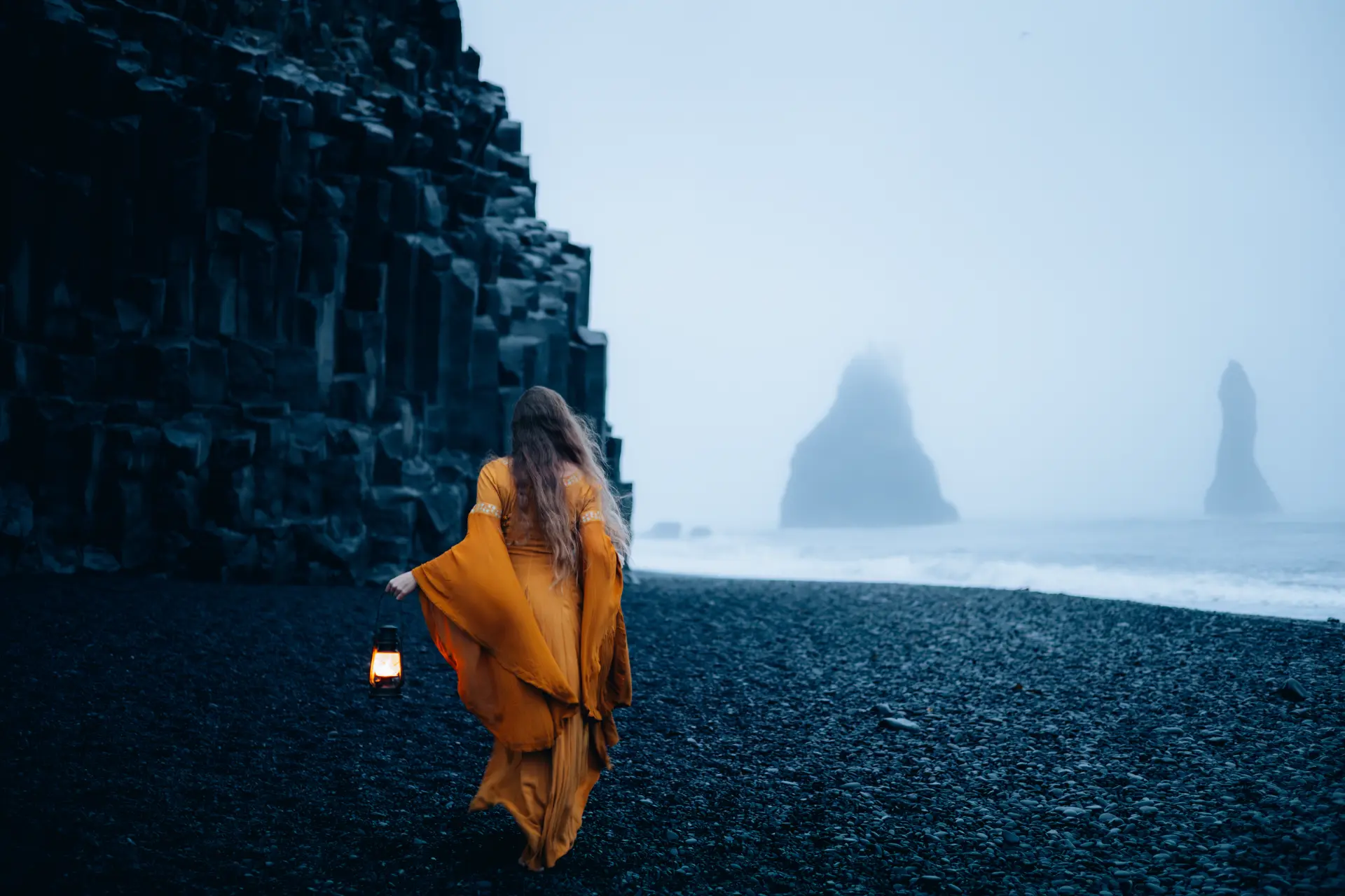 Woman in an amber dress carrying a lantern on a black sand beach