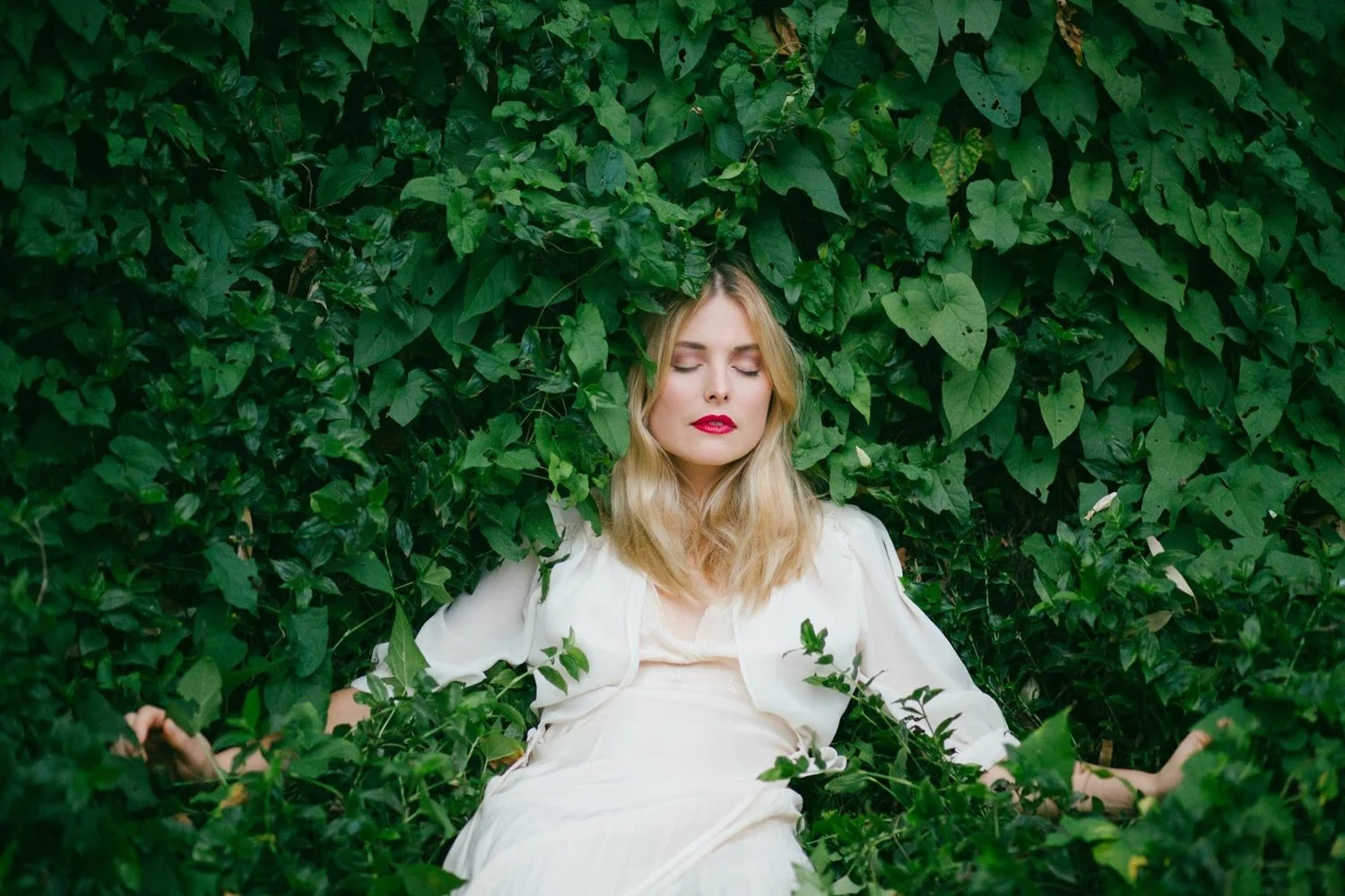 Blonde woman in a white dress resting in a wall of green leaves