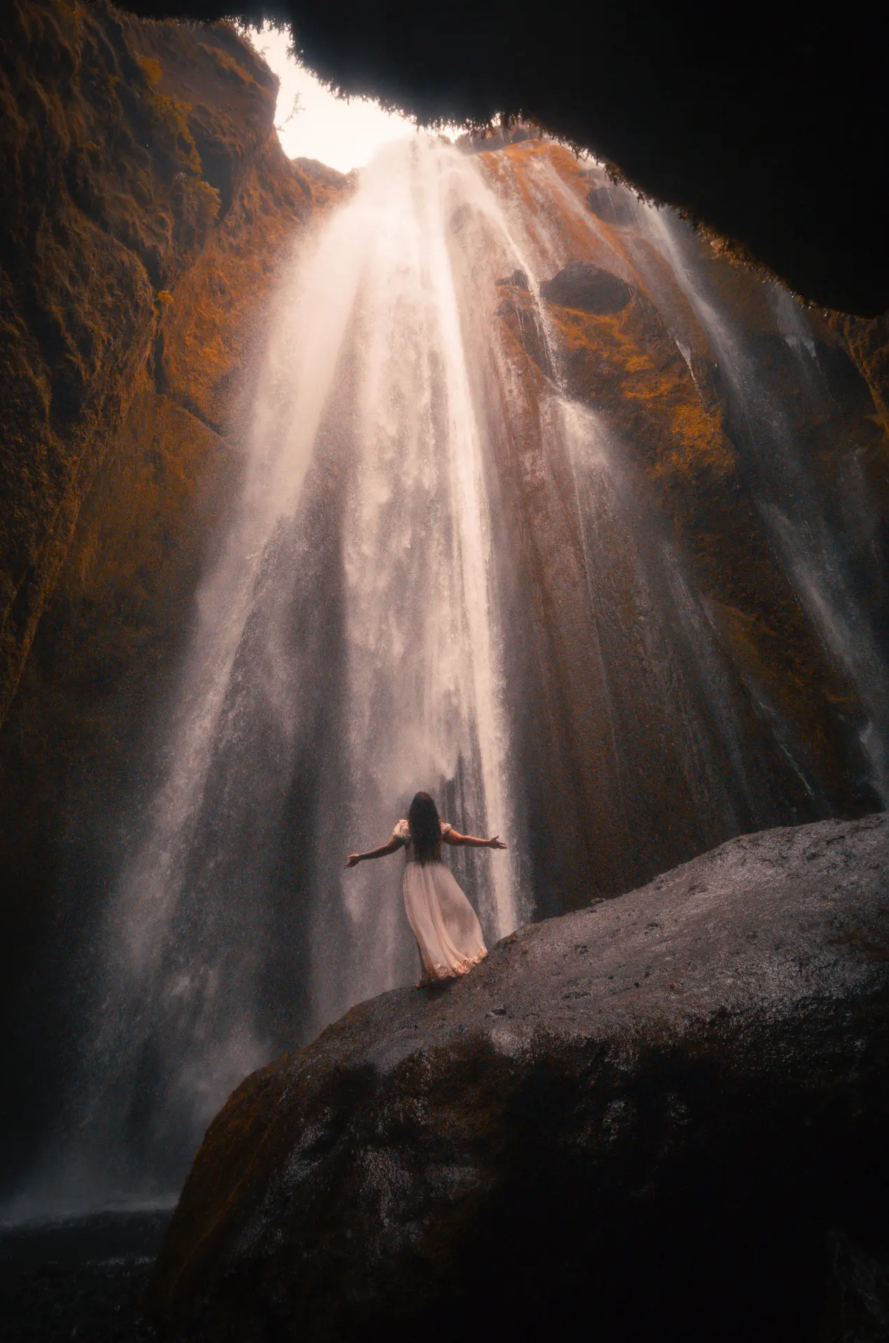 Woman in a pale gown beneath a sunlit waterfall