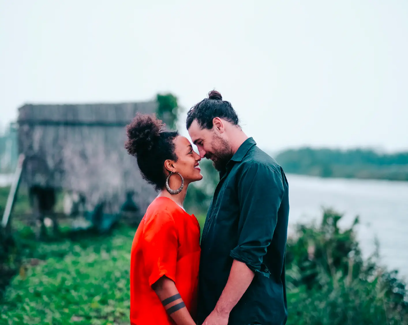 Couple in red and green touching foreheads by a riverside hut