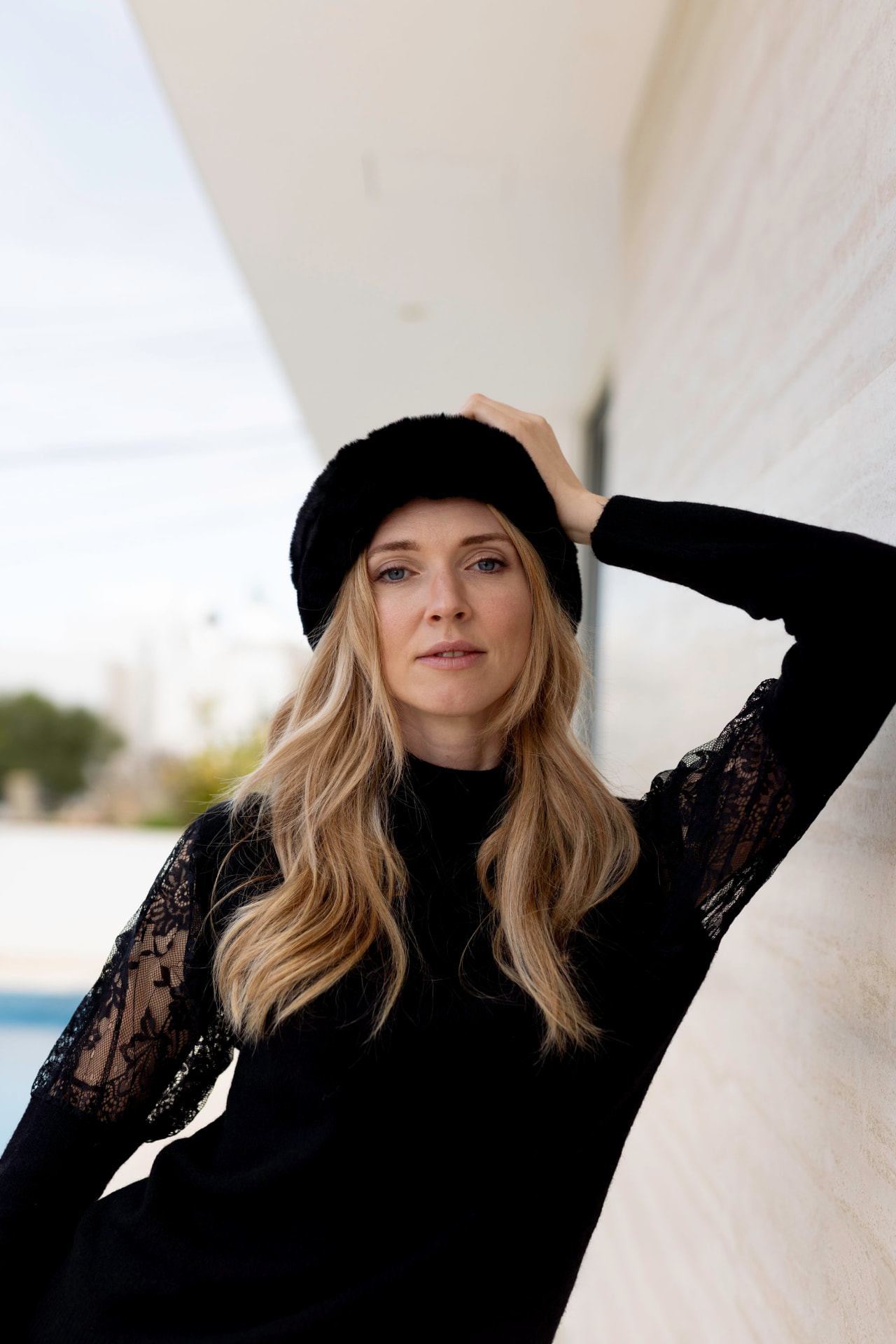 Blonde model in a black lace dress and fur hat against a white wall