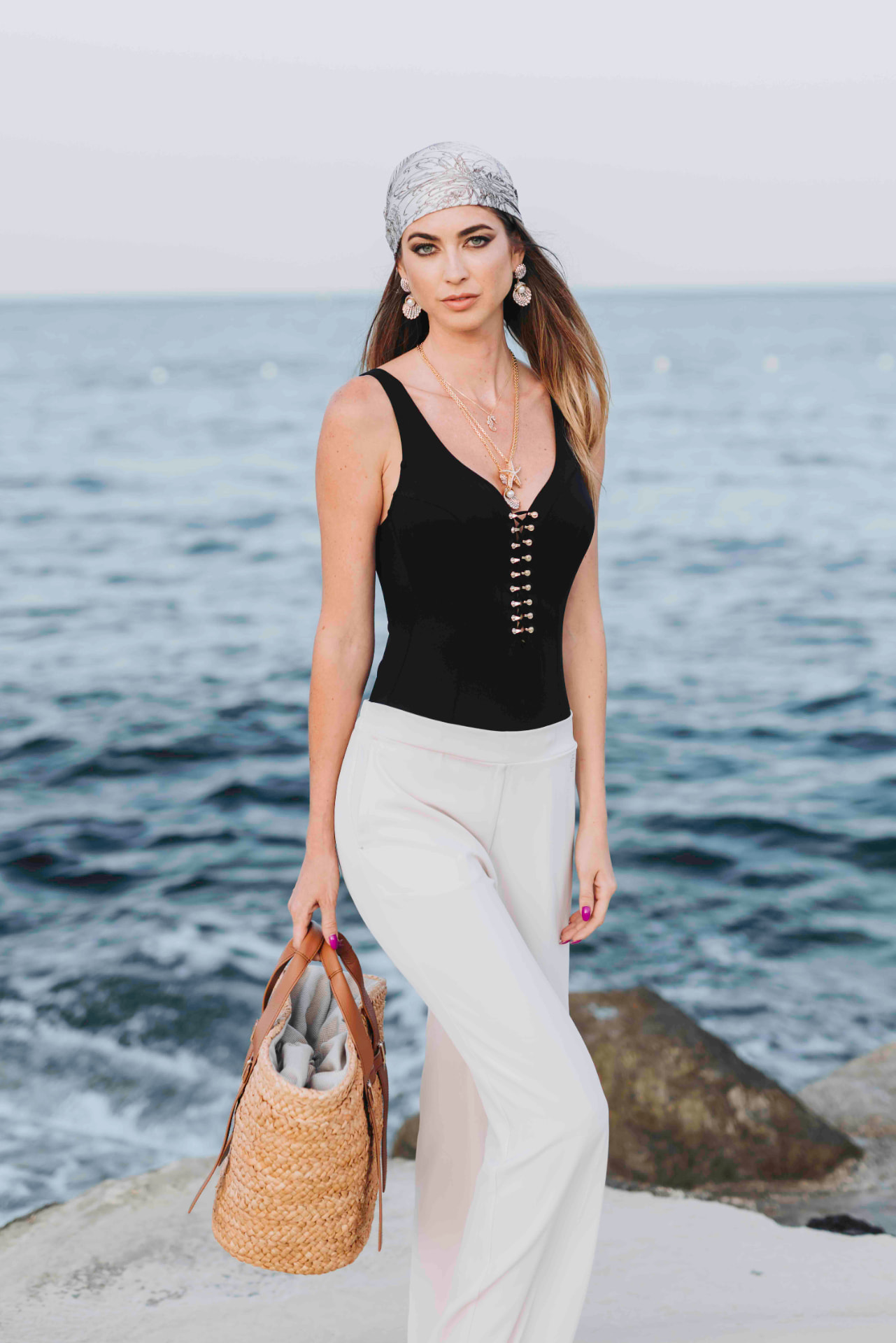 Model in a black bodysuit and white trousers with a straw tote by the sea