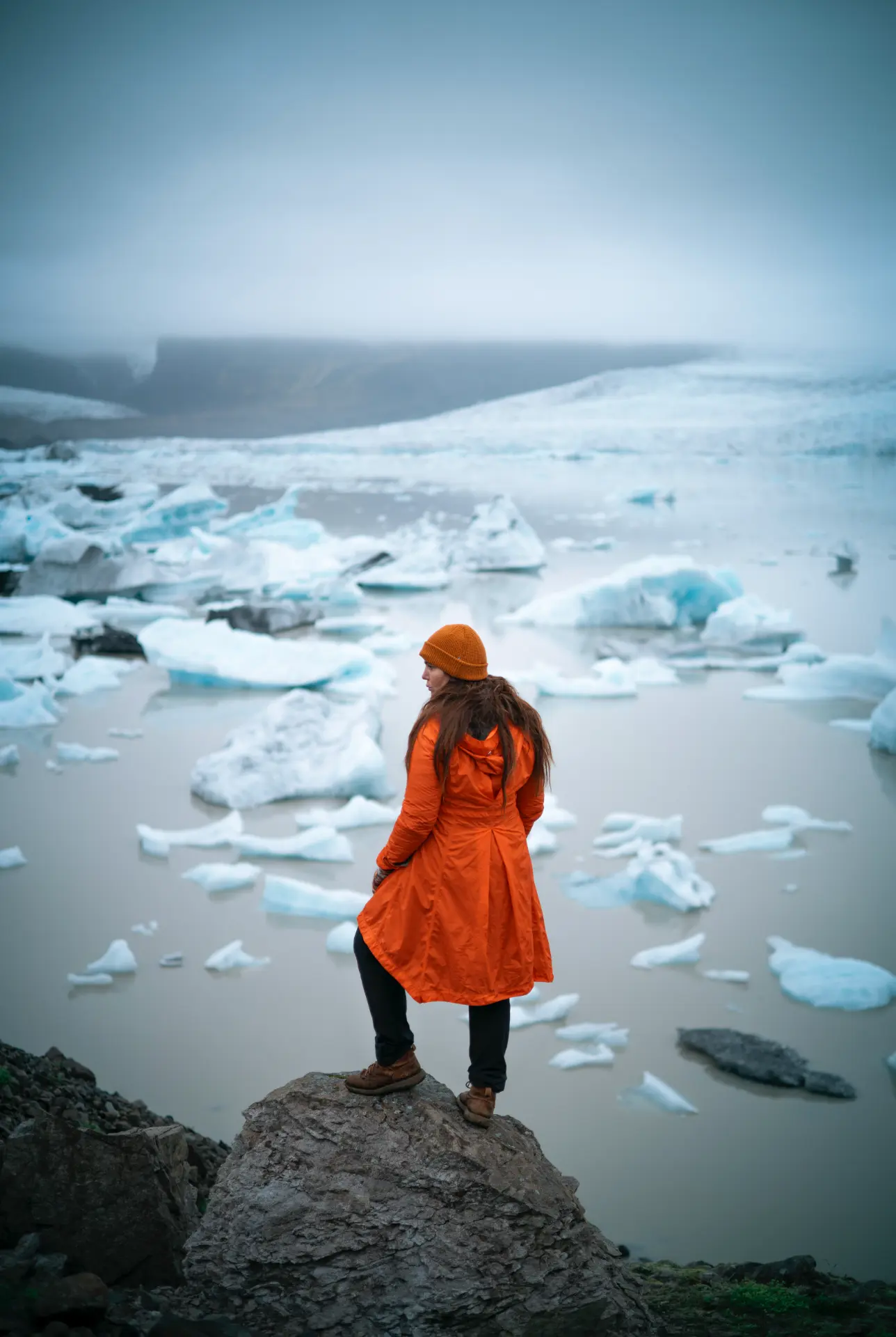 Woman in an orange coat on a rock above a glacier lagoon