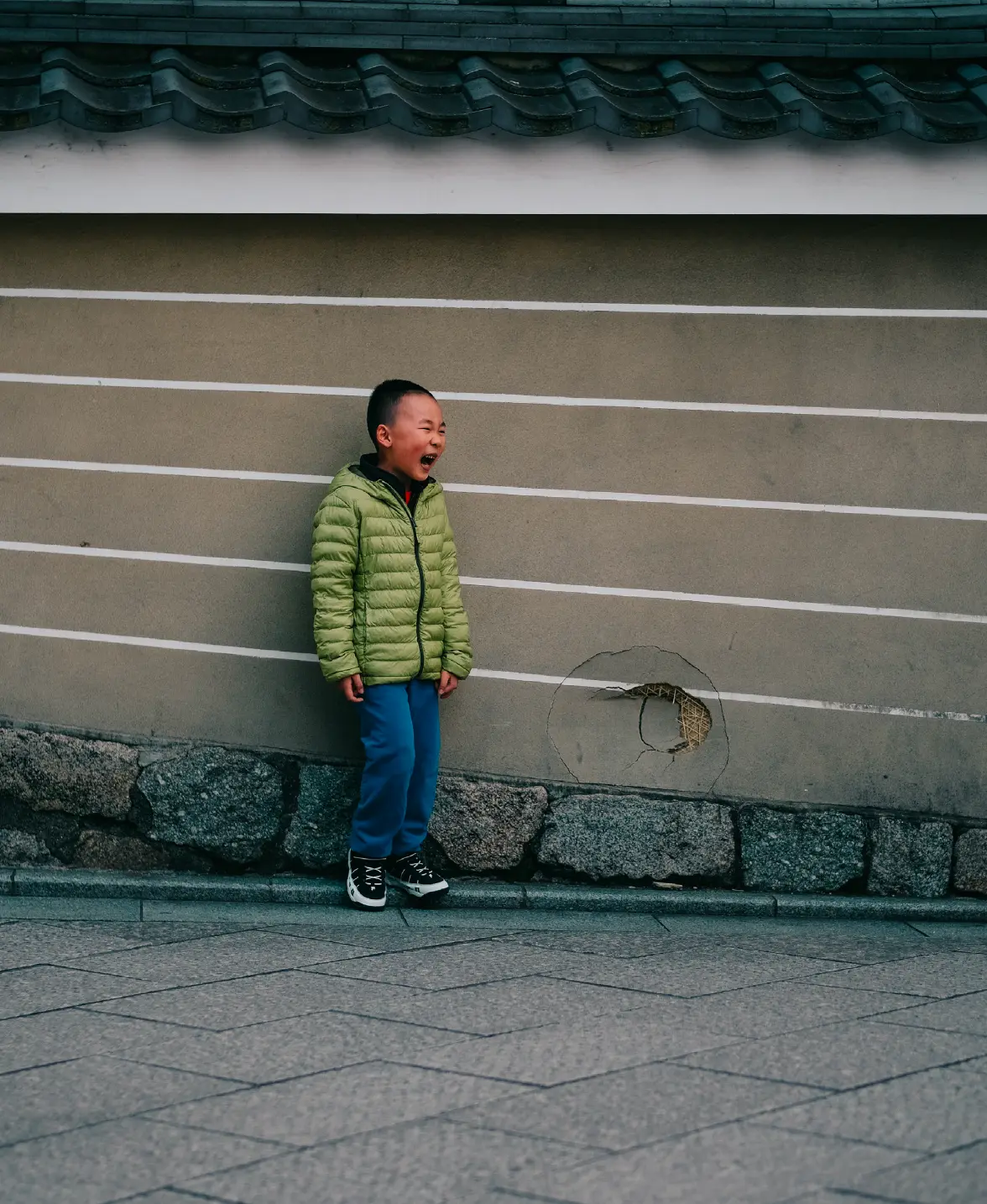 Boy in a green jacket laughing against a striped temple wall in Japan