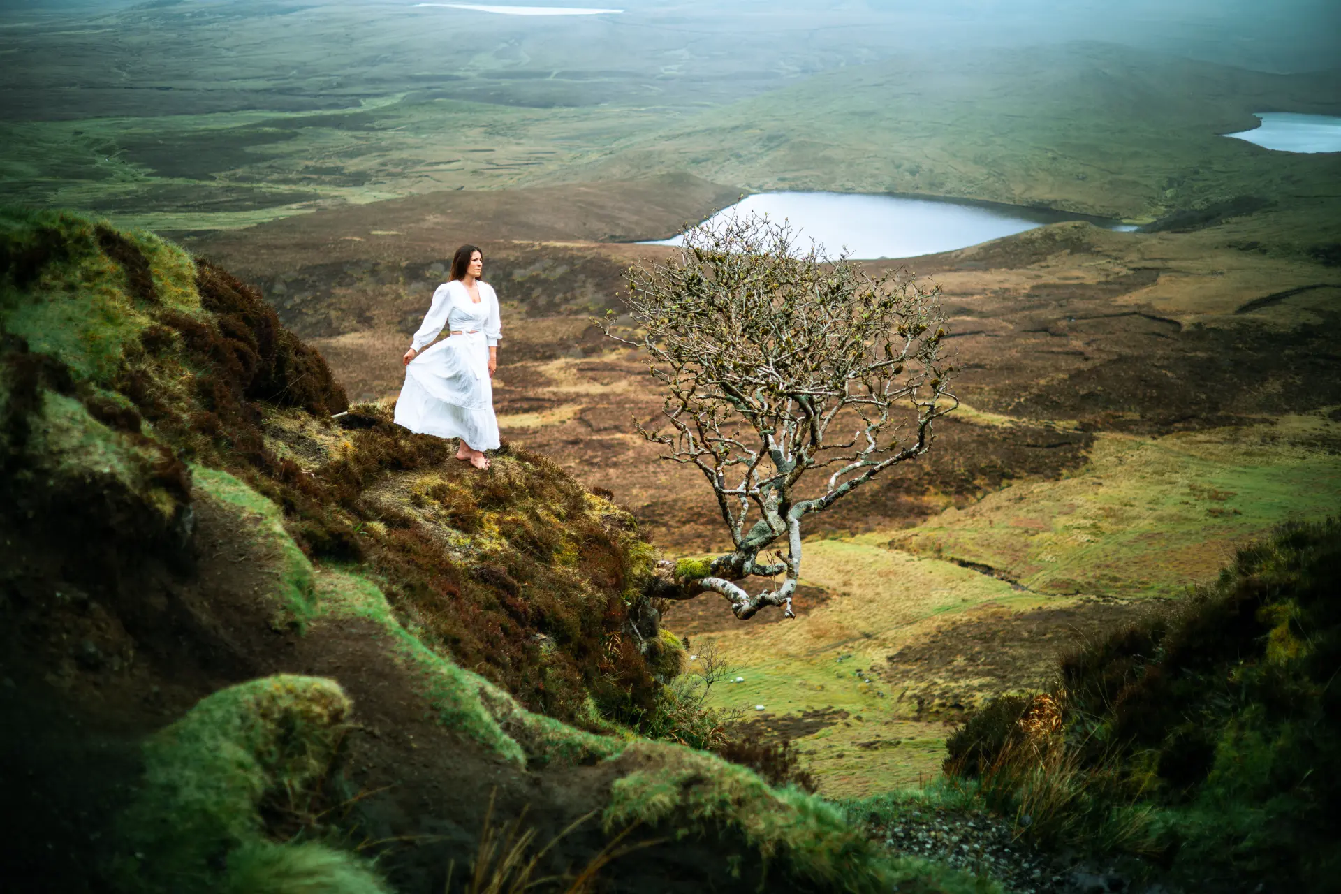 Woman in a white dress on a mossy ridge beside a lone windswept tree