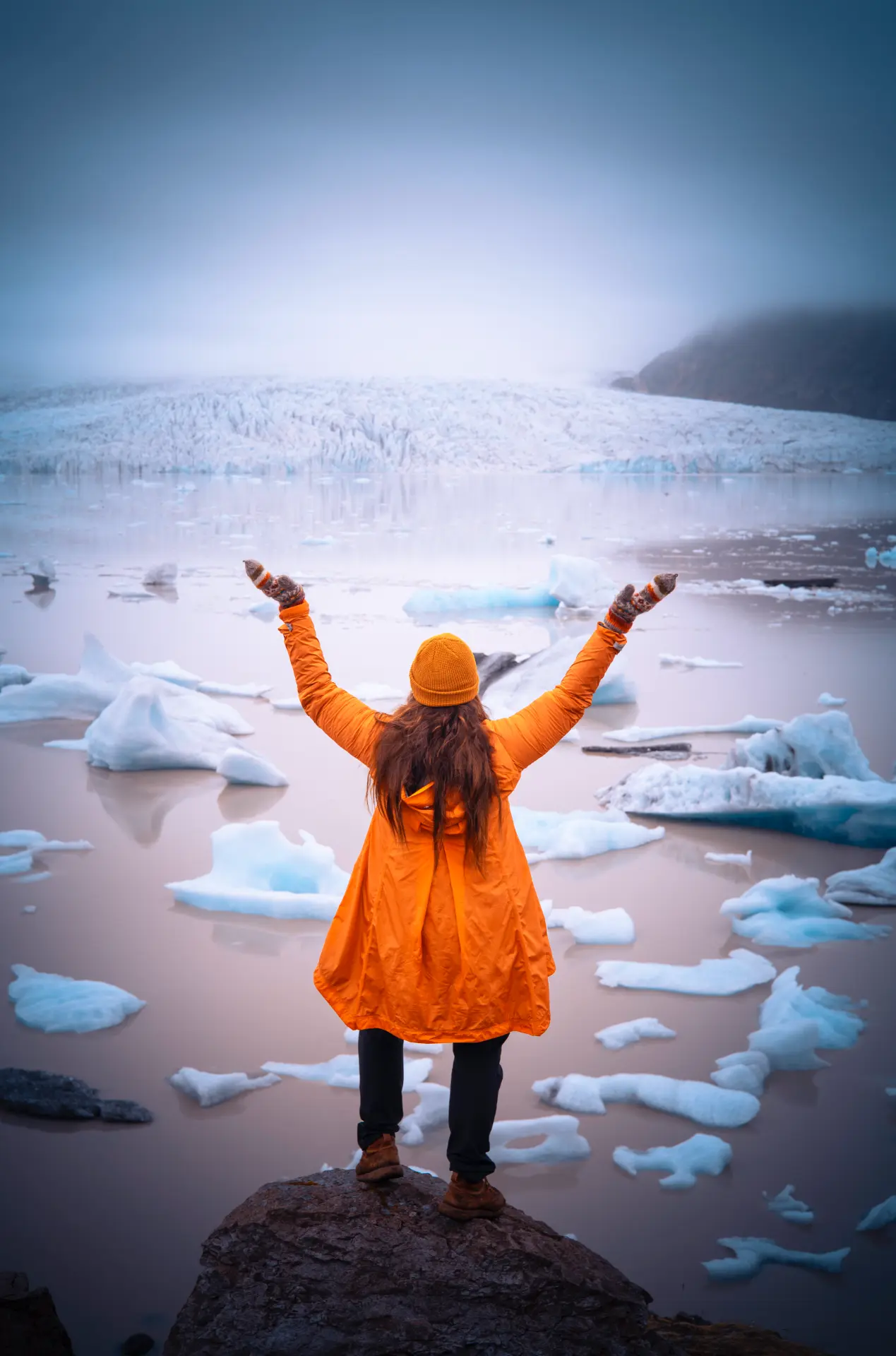 Woman in an orange coat with arms raised before a glacier