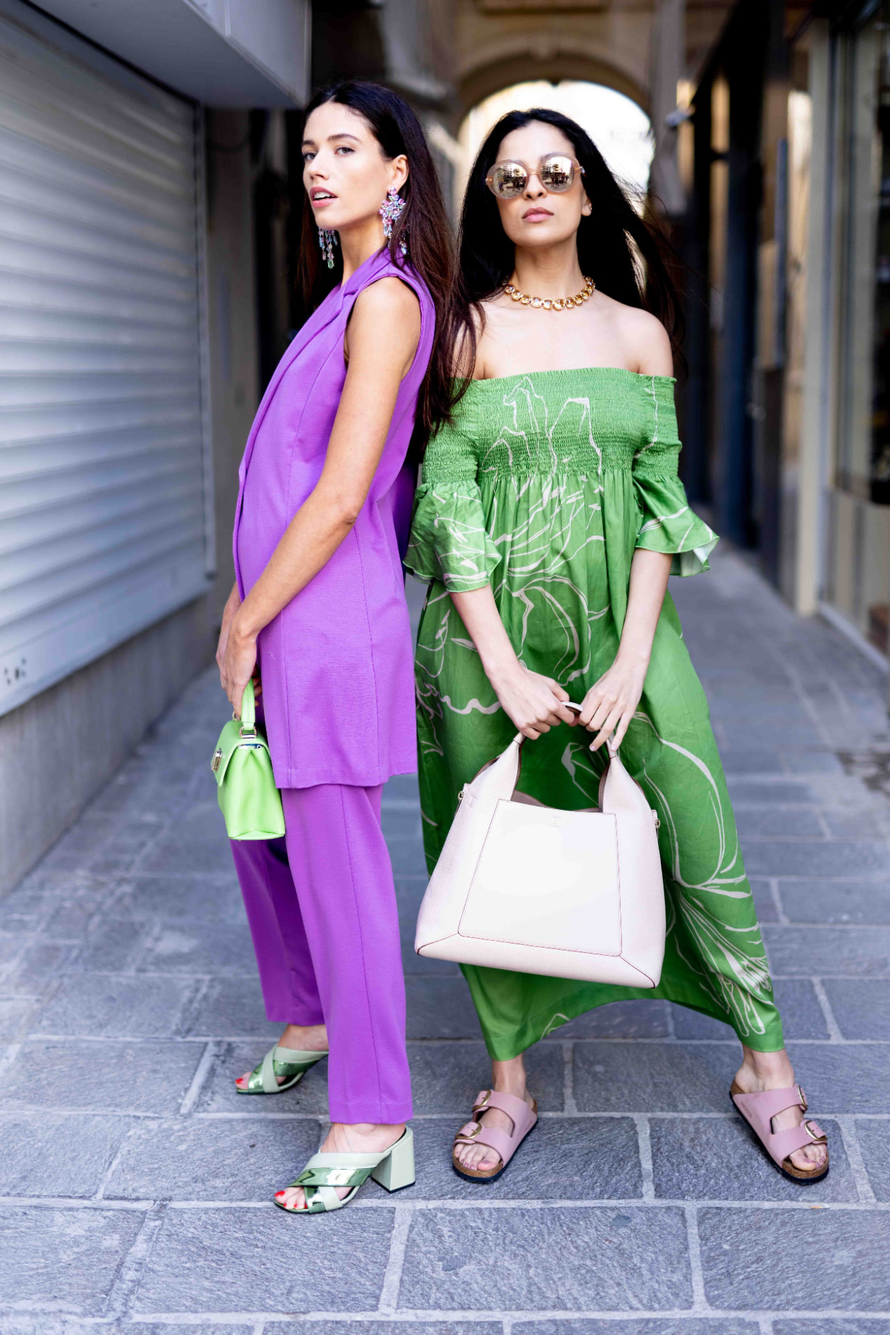 Two models in purple and green outfits with designer bags on a street