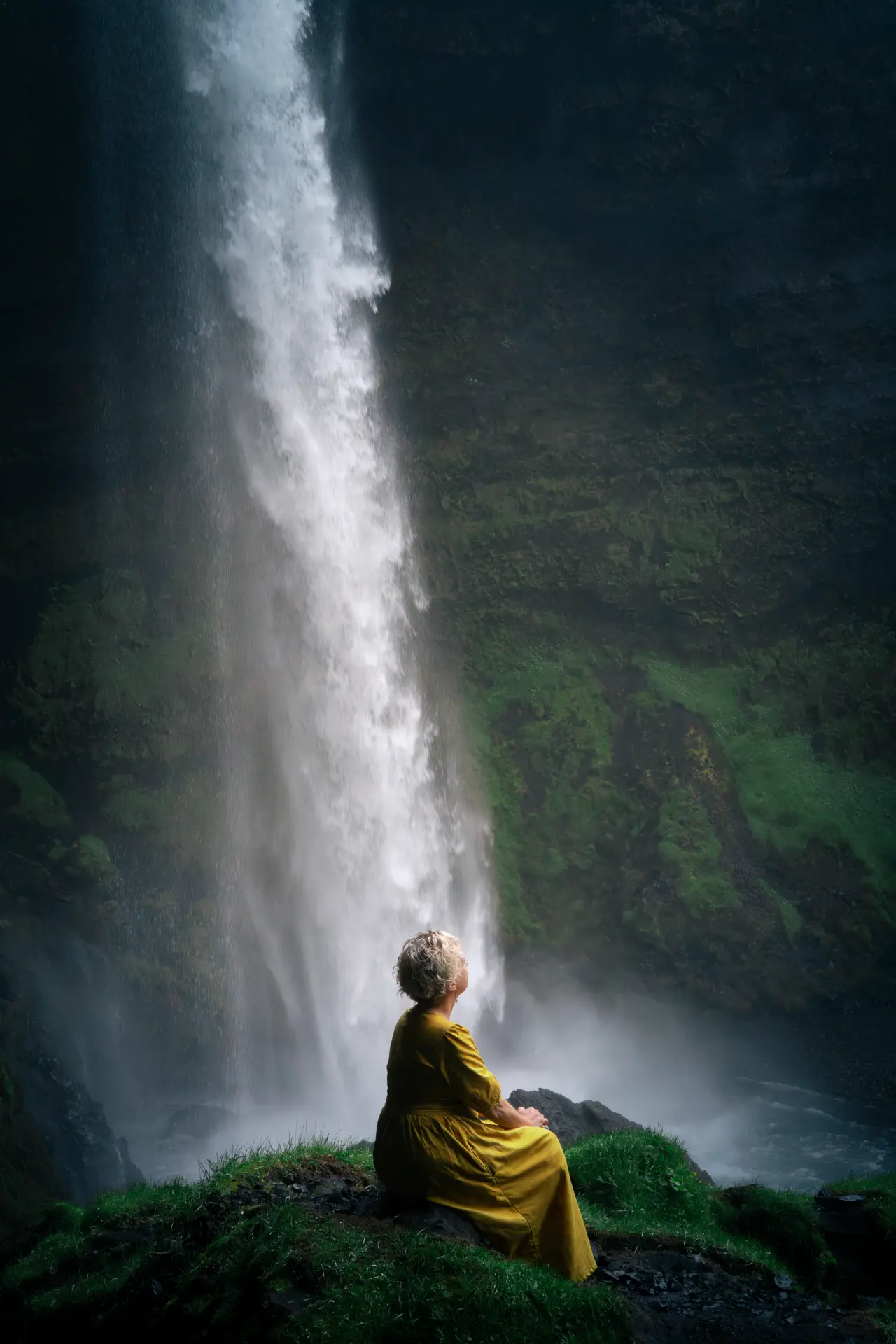 Woman in a gold dress seated beneath a tall waterfall