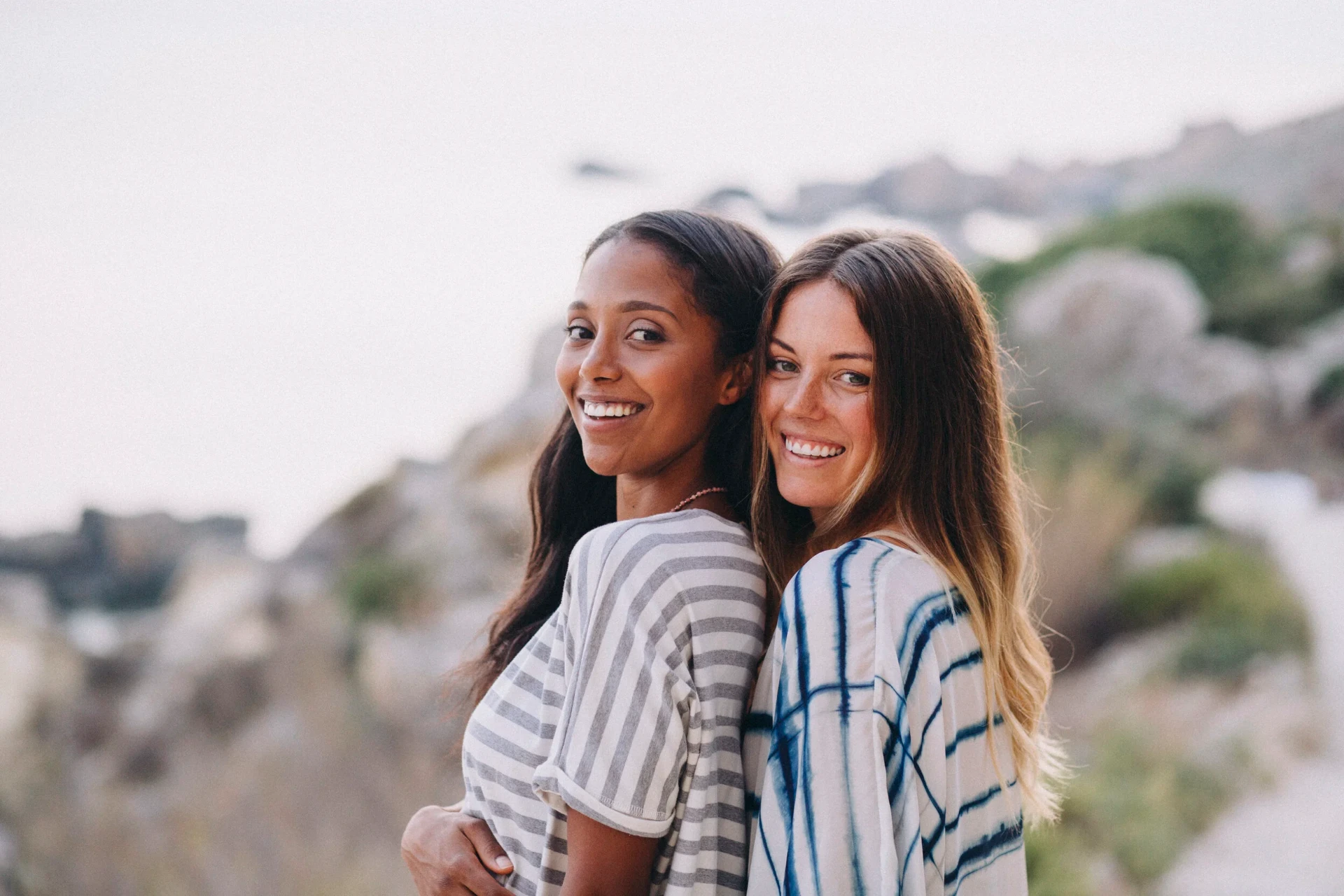 Two models smiling back to back above a rocky bay