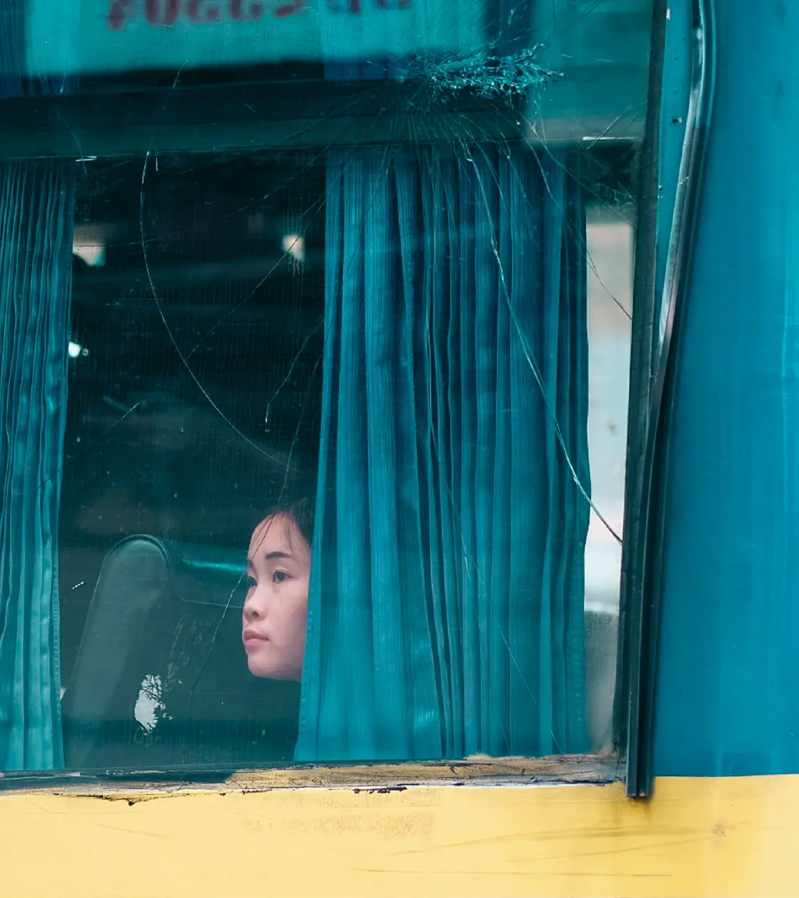 Woman's face framed by teal curtains behind a cracked bus window