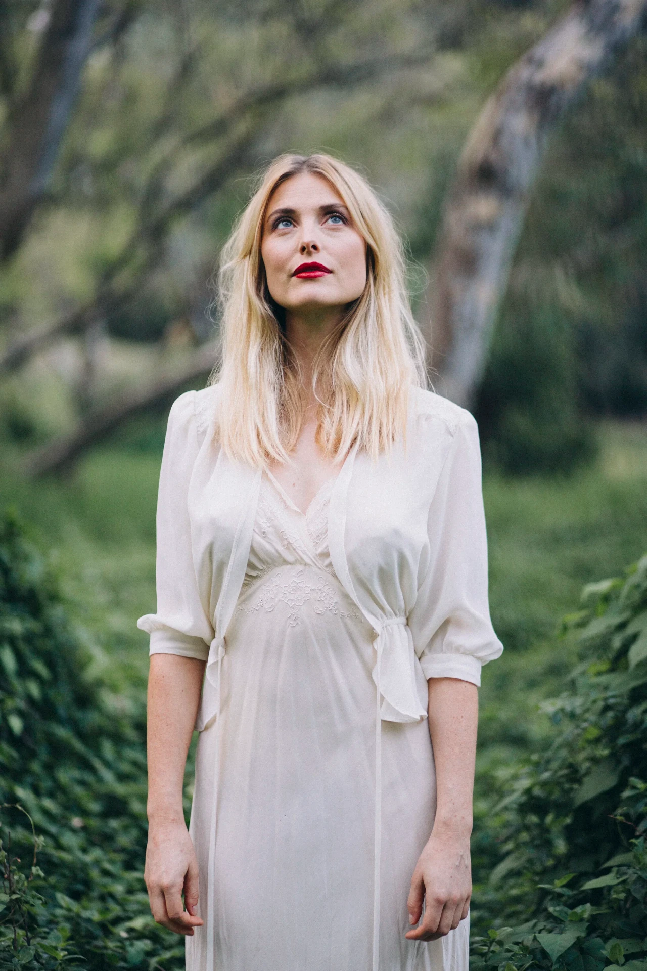 Blonde woman in a white vintage dress looking upward in woodland
