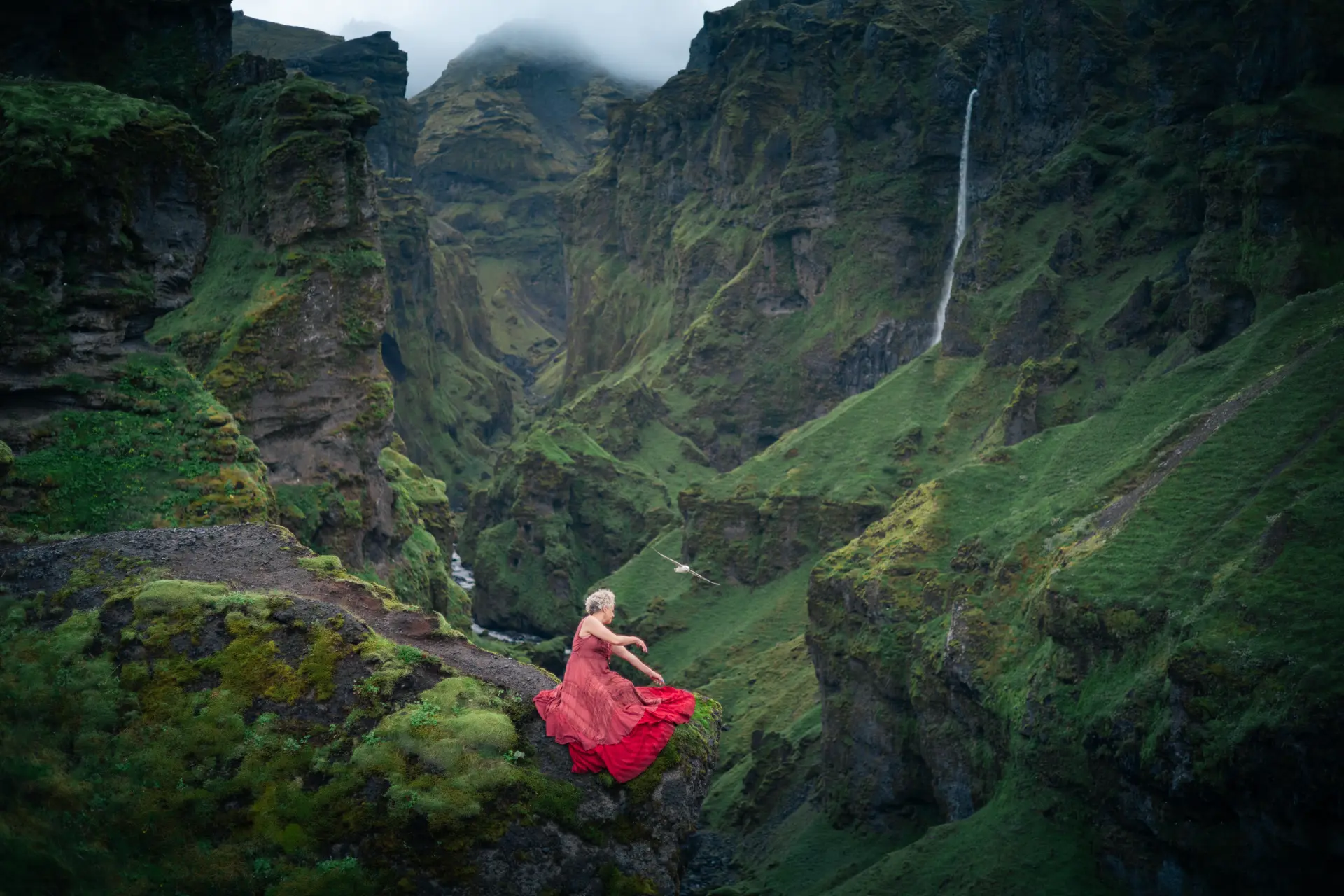 Woman in red seated above a moss-green Icelandic canyon
