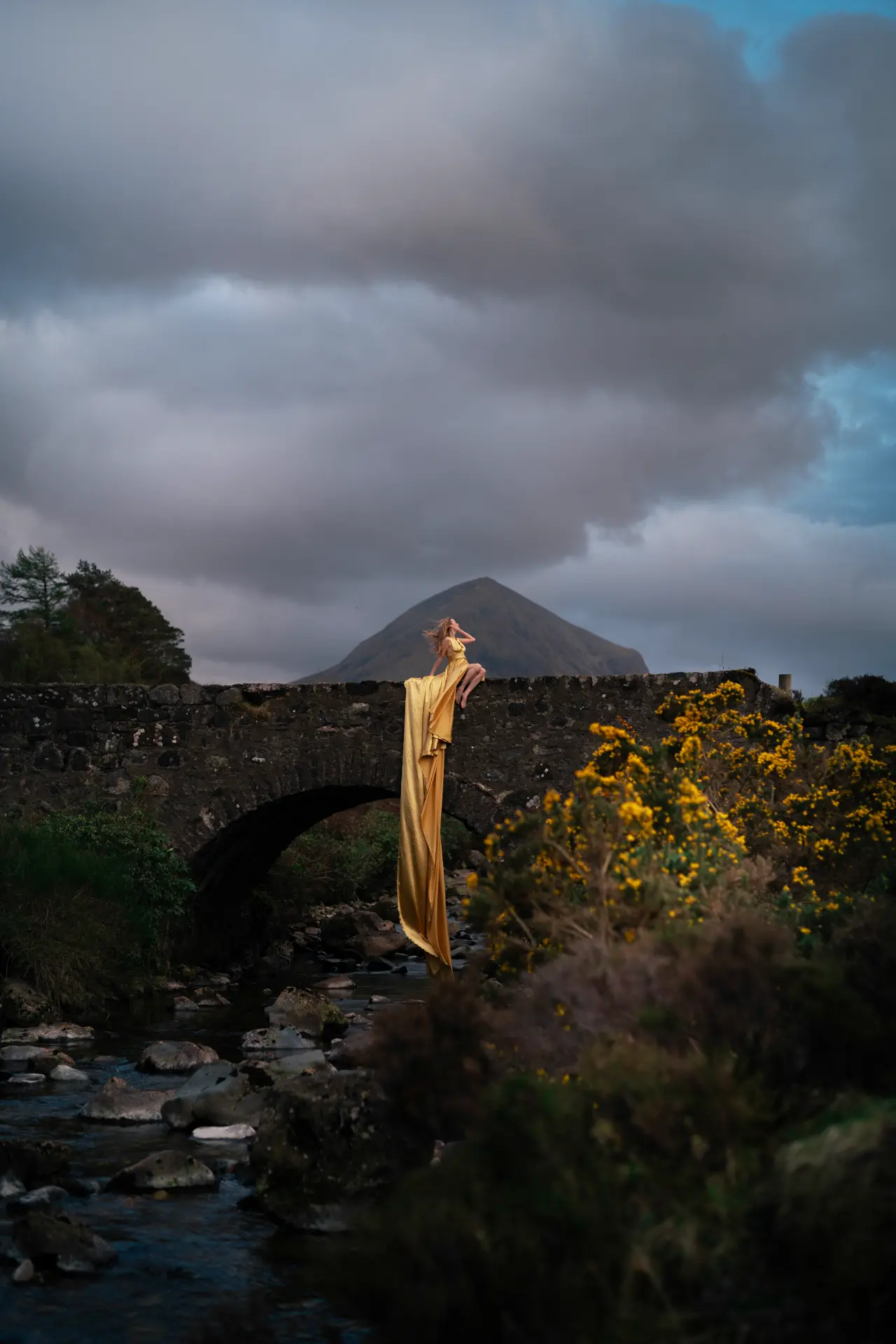 Woman in a gold gown on the Sligachan bridge beneath a stormy sky