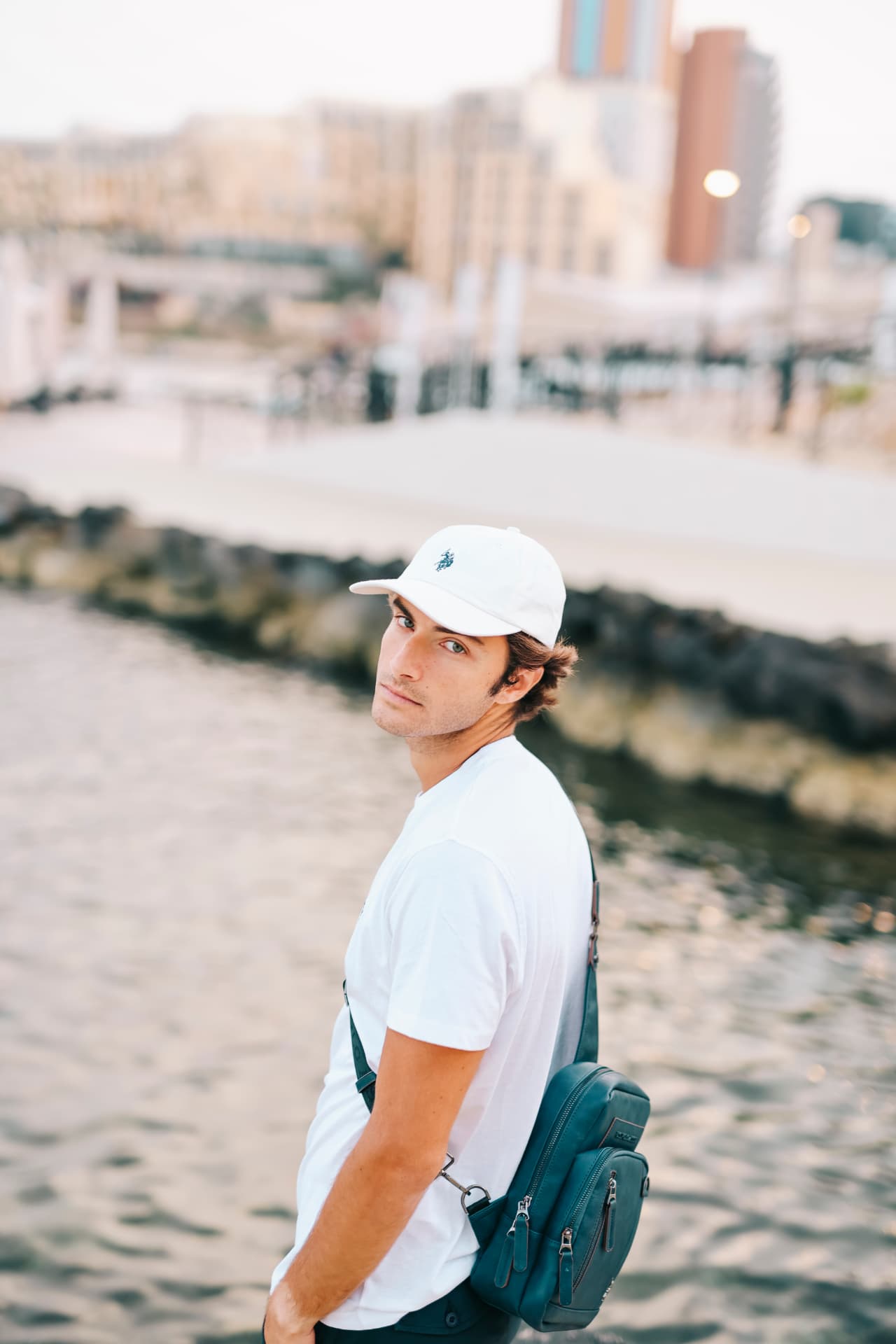 Man in a white cap and tee with a leather sling bag by the waterfront