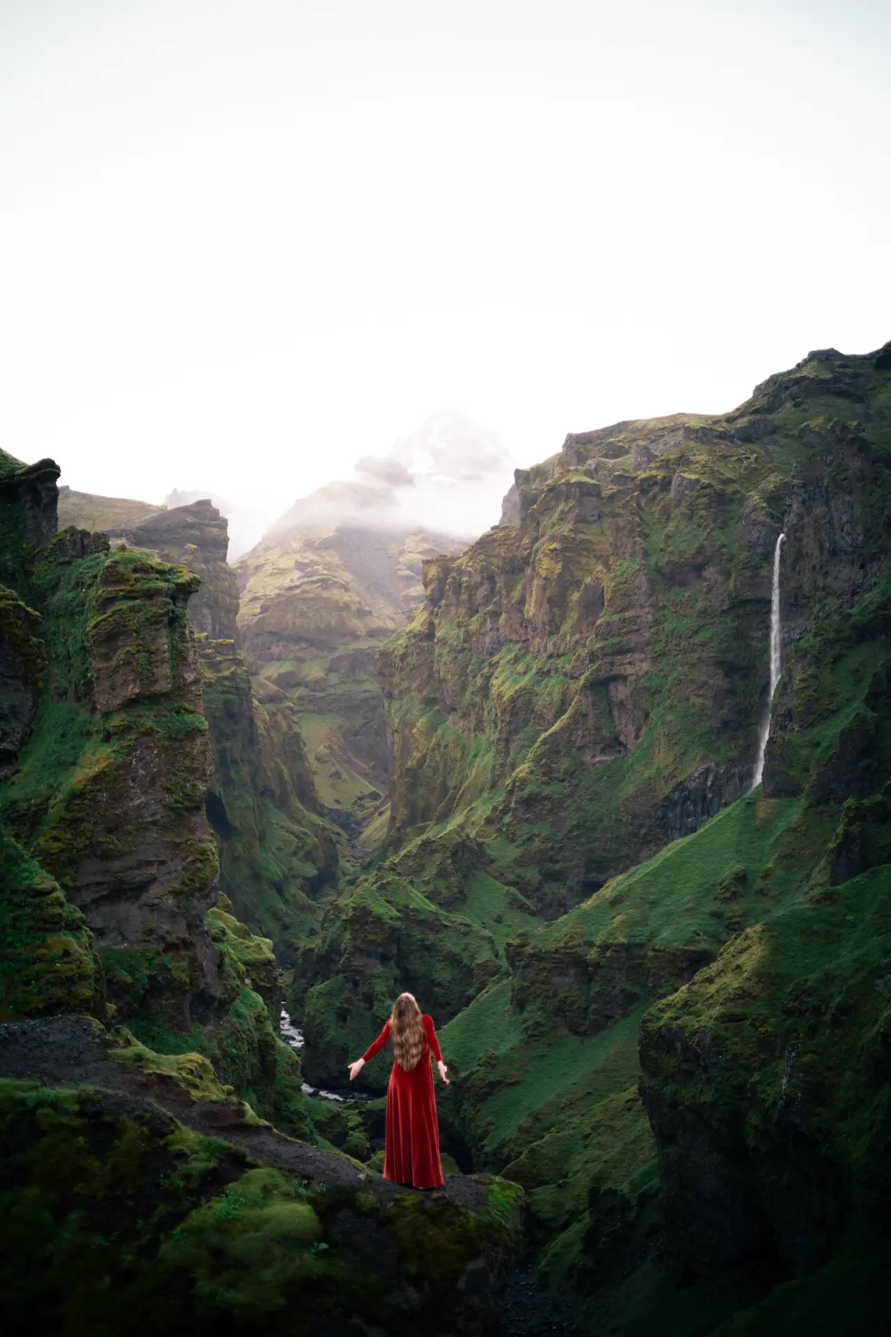 Woman in a red velvet dress facing a mossy canyon and waterfall