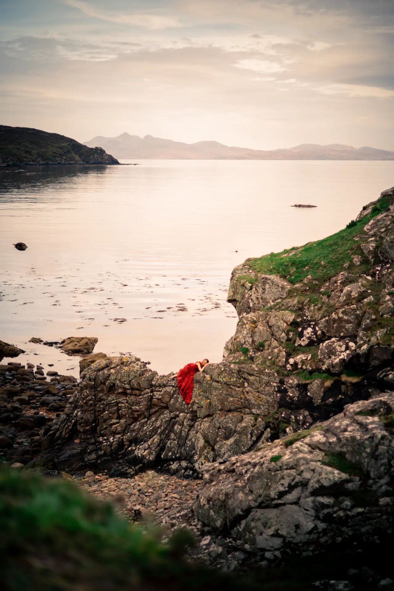 Figure in a red gown resting on coastal rocks at golden hour